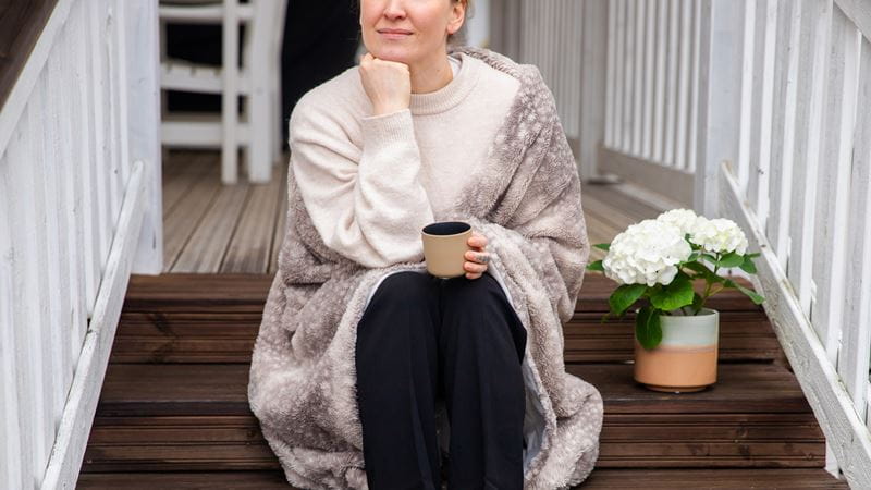 Sp-Kodin brändikuva- Nainen istuu omakotitalon ulkorappusilla kahvi kädessä ja viltti päällä.